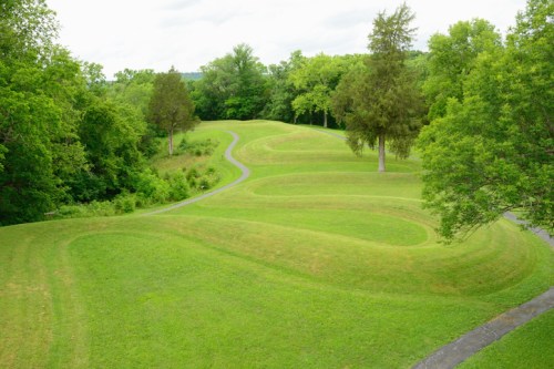 A lush green landscape with rolling hills and winding paths, surrounded by trees under a cloudy sky.