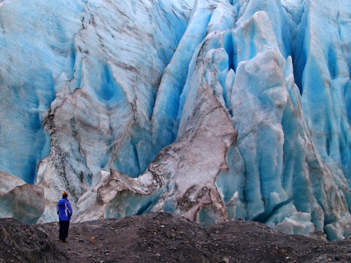 A person stands in front of a massive blue glacier, surrounded by rocky terrain and ice formations.