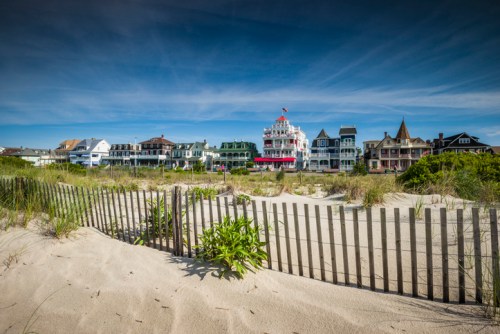 A sandy beach with a wooden fence, featuring colorful Victorian-style houses in the background under a blue sky.