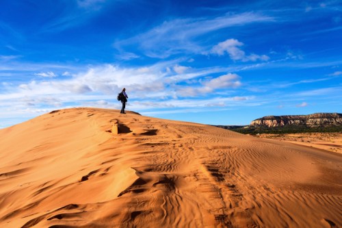 A lone hiker stands atop a sandy dune under a blue sky with wispy clouds, surrounded by desert landscape.