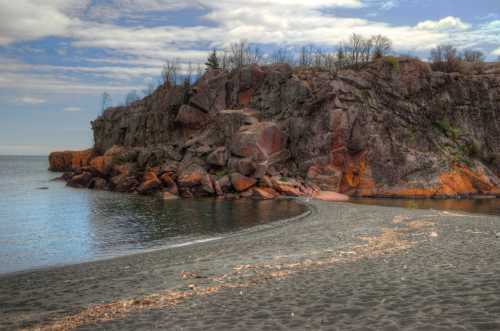 A rocky shoreline with a sandy beach, calm water, and a cloudy sky in the background.