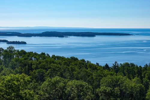 A serene view of a calm blue lake surrounded by lush green trees and distant islands under a clear sky.