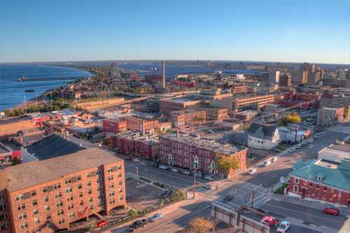 Aerial view of a coastal city with buildings, roads, and a harbor under a clear blue sky.