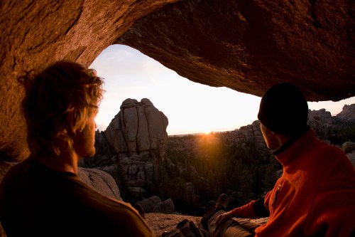 Two people sit in a rock cave, watching a sunset over a mountainous landscape.