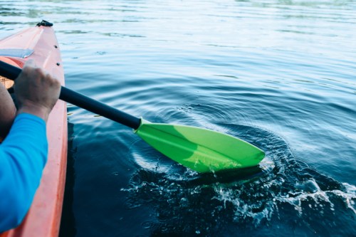 A close-up of a hand holding a green kayak paddle, splashing water as it moves through calm lake waters.