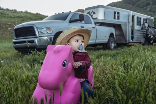 A baby in a cowboy hat rides a pink toy horse in a grassy field, with a truck and horse trailer in the background.