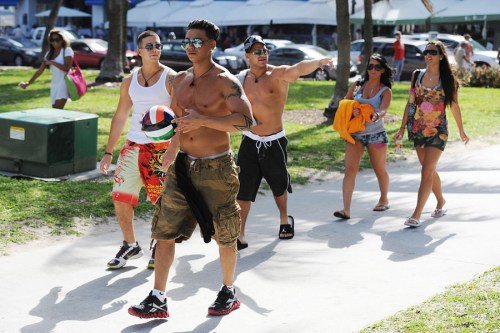 A group of young adults walking on a sunny path, some holding a volleyball, dressed in summer attire.