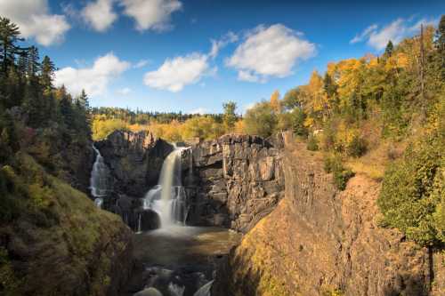 A scenic waterfall cascading over rocky cliffs, surrounded by vibrant autumn foliage and a clear blue sky.