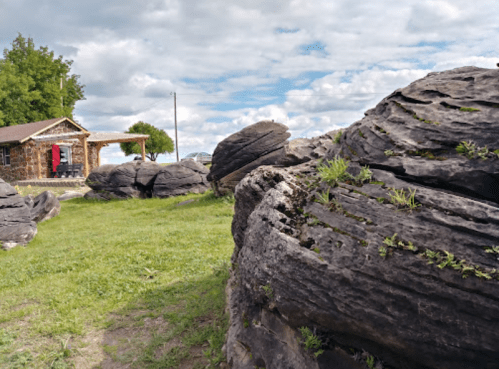 A grassy area with large, textured rocks and a rustic cabin in the background under a cloudy sky.