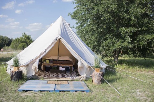 A spacious white tent set up on grass, featuring a wooden bed and decorative elements, surrounded by trees.