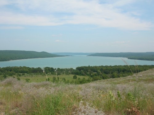A serene view of a lake surrounded by green hills and fields under a blue sky with scattered clouds.