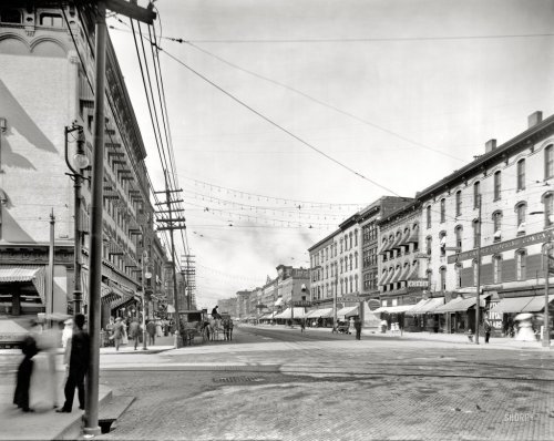 Historic street scene featuring buildings, streetcars, and pedestrians, with overhead wires and awnings.