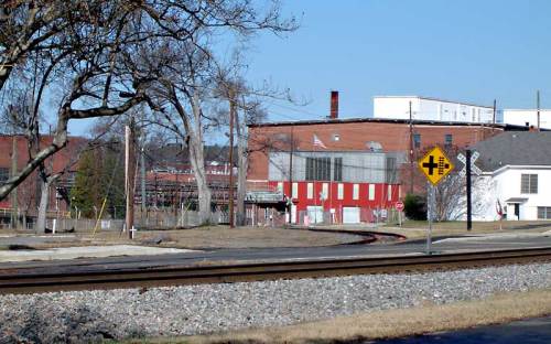 A view of a railway crossing sign near a factory, with trees and a clear blue sky in the background.