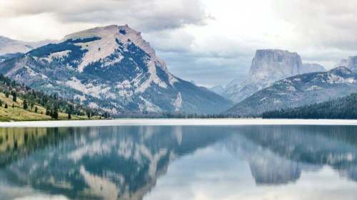 A serene lake reflects mountains under a cloudy sky, surrounded by lush greenery.