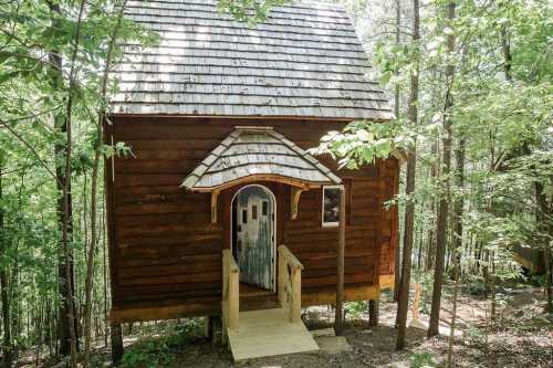 A small wooden cabin with a sloped roof, surrounded by trees and a wooden walkway leading to the entrance.