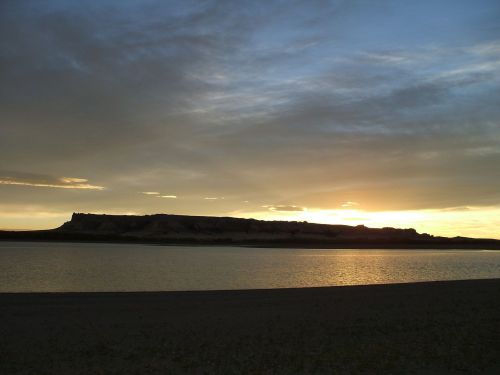 Silhouette of a rocky landscape at sunset, reflecting on a calm body of water under a cloudy sky.