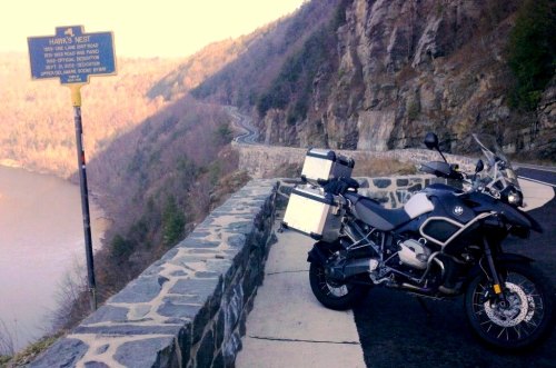 A motorcycle parked by a scenic overlook with a river and mountains in the background, near a historical sign.