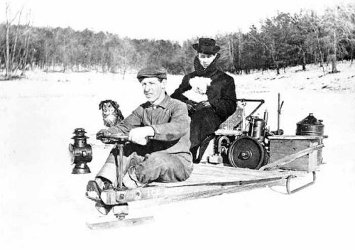 Two people and a dog on a wooden sled on ice, with a vintage engine and equipment in the background.
