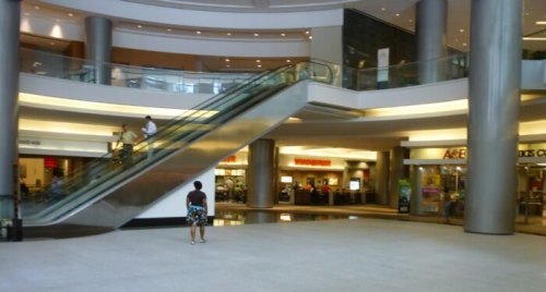 A spacious mall interior featuring an escalator, shops in the background, and a person standing on the floor.