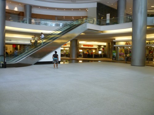 A spacious mall interior with an escalator, a person standing alone, and shops in the background.