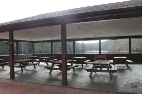 A covered picnic area with wooden tables overlooking a calm lake and wooded landscape in the background.