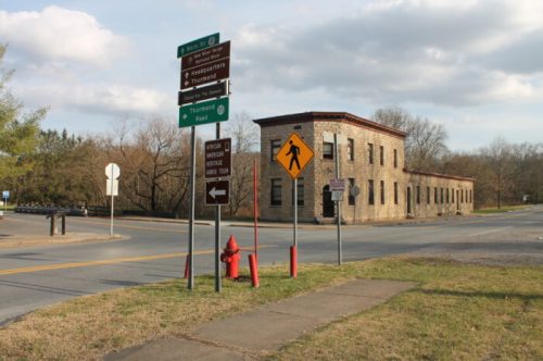 A stone building at a road intersection with directional signs and a pedestrian crossing warning.