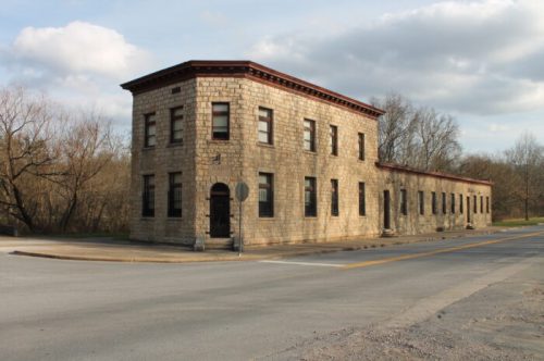 Historic stone building at a street corner, featuring large windows and a flat roof, surrounded by bare trees.
