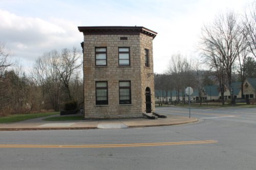 A small, historic stone building at a street corner, surrounded by trees and a few other structures in the background.