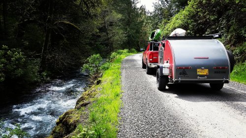 A red vehicle with a trailer parked beside a winding gravel road next to a flowing stream, surrounded by lush greenery.