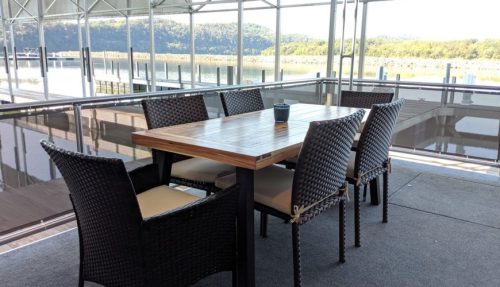 A wooden dining table with six wicker chairs on a deck overlooking a calm water view.