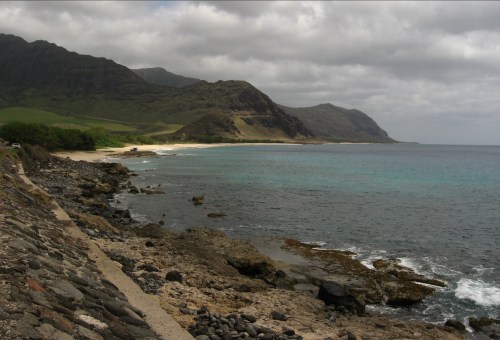 A rocky coastline with turquoise waters, mountains in the background, and a cloudy sky above.