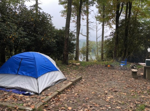 A blue and gray tent set up in a wooded campsite near a lake, with a fire pit and chairs nearby.