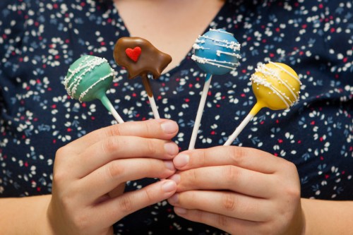 A person holds four colorful cake pops on sticks, decorated with icing and sprinkles, against a floral background.