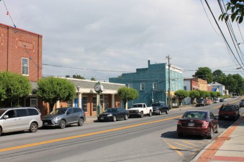 A small town street lined with trees, shops, and parked cars under a cloudy sky.