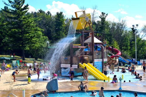 A vibrant water park scene with children playing, a large water slide, and a splash pad under a sunny sky.