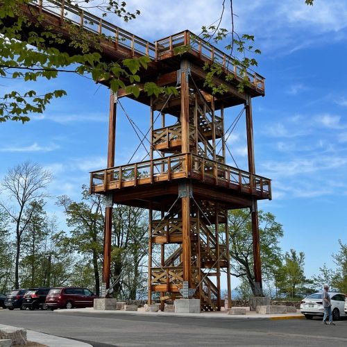 A wooden observation tower surrounded by trees, with a clear blue sky in the background and parked cars nearby.
