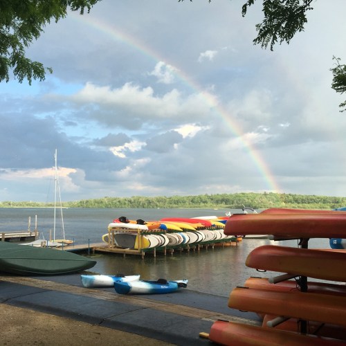 A serene lakeside view with colorful kayaks, a rainbow in the sky, and boats docked along the shore.