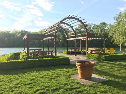 A wooden pergola with seating overlooks a serene lake, surrounded by lush greenery and a large flower pot.