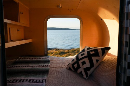 Cozy interior of a camper with a view of a serene lake and distant hills, featuring a patterned pillow and blankets.