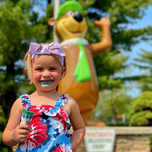 A young girl with blue ice cream on her face smiles, holding an ice cream cone in front of a cartoon bear statue.