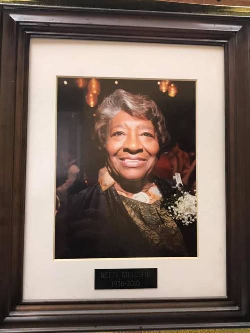 Framed portrait of a smiling woman with curly hair, wearing a black and gold outfit, honoring her memory.