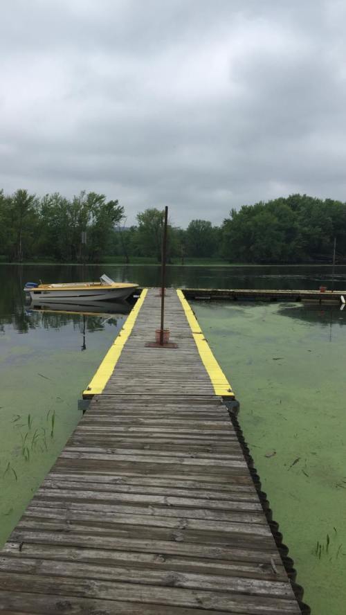 A wooden dock extends into a calm, green-tinted lake, with a small boat moored nearby under a cloudy sky.