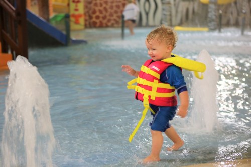 A smiling child in a life jacket walks through a shallow pool, splashing water around in a playful setting.