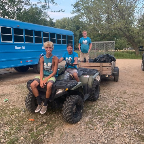 Two young men sit on an ATV, with a blue bus and a man in the background. They are outdoors in a gravel area.