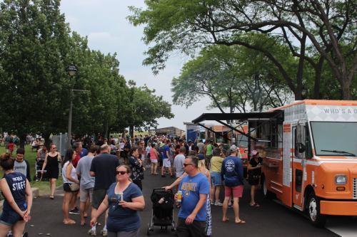 A busy outdoor food festival with crowds, food trucks, and trees in the background on a cloudy day.