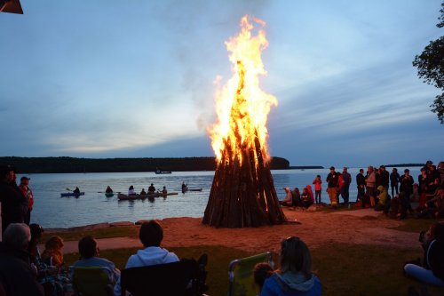 A large bonfire burns brightly by the water, with people gathered around and kayakers in the background at dusk.