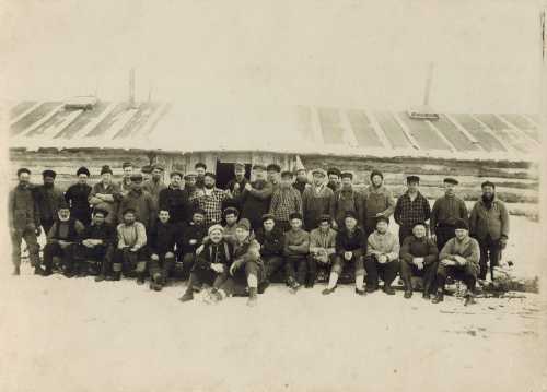 A large group of men poses in front of a log building in a snowy landscape, dressed in winter clothing.