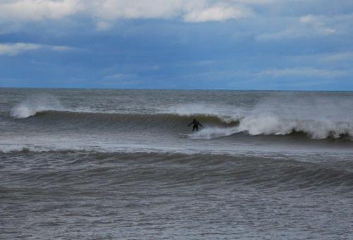 A lone surfer rides a wave under a cloudy sky, with gentle waves rolling in from the ocean.
