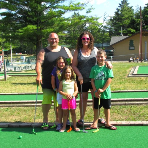 A family of five stands on a mini-golf course, smiling and holding putters, surrounded by trees and a colorful backdrop.