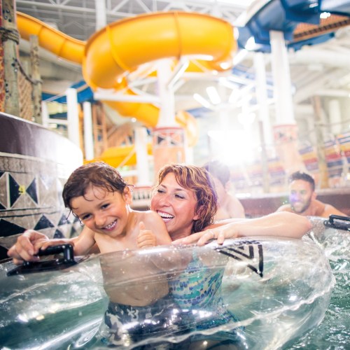 A woman and a boy smile while floating on a tube in a water park, with a water slide in the background.